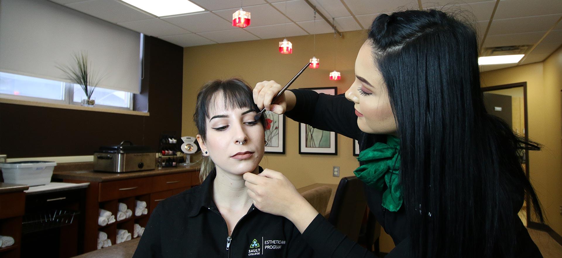 One female Esthetician student applies make-up to another Esthetician student in the о salon spa. 